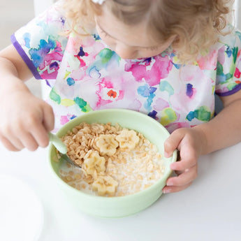 A child in a colorful floral bib enjoys cereal with banana slices from a Bumkins Silicone Grip Bowl: Sage at a white table.