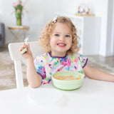 A curly-haired child beams, enjoying cereal from a Bumkins Silicone Grip Bowl: Sage, adding charm with their bright bib and bow.