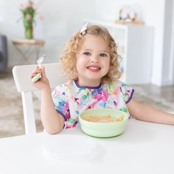 A curly-haired child beams, enjoying cereal from a Bumkins Silicone Grip Bowl: Sage, adding charm with their bright bib and bow.