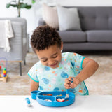 A curly-haired child enjoys toddler portions from a dark blue silicone dish by Bumkins, wearing a colorful bib in a blurred living room.