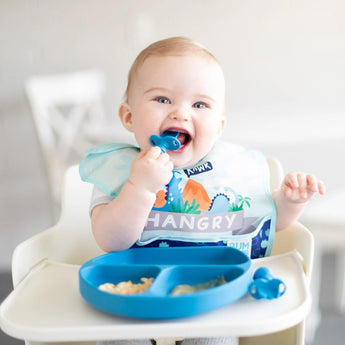 A smiling baby in a high chair wears a HANGRY dinosaur bib holding a spoon with food on a Bumkins Silicone Grip Dish: Dark Blue.