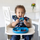 A cheerful toddler in a high chair with food on a Bumkins Silicone Grip Dish in Dark Blue, wearing a bib, holding a spoon.