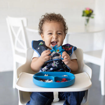 A cheerful toddler in a high chair with food on a Bumkins Silicone Grip Dish in Dark Blue, wearing a bib, holding a spoon.