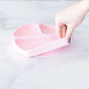 A hand lifts a Bumkins Silicone Grip Dish: Pink from a marble countertop.