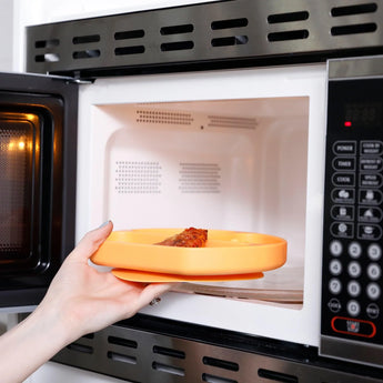 A hand places the Bumkins Silicone Grip Dish: Tangerine into the open microwave, revealing its cozy interior.
