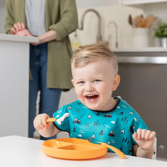 A toddler with a Bumkins tangerine Silicone Grip Dish and blue bib holds a fork as an adult stands by the counter.