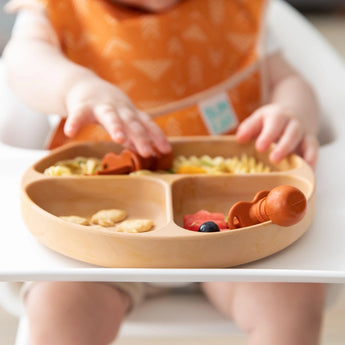 Close-up of a baby in a high chair reaching for toddler utensils in a Bumkins Silicone Grip Dish: Wood Grain filled with food.