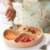 Close-up of a child eating spaghetti from a Bumkins Silicone Grip Dish: Wood Grain with a fork at a white table, wearing a colorful bib.