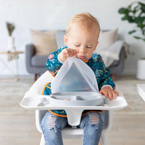 A toddler in a high chair wearing a colorful bib eagerly opens a Bumkins Silicone Grip Dish with Lid (3 Section): Gray in a cozy living room.