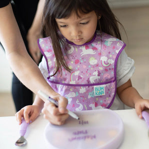An adult writes on the lid of a Bumkins Silicone Grip Dish with Lid (3 Section): Lavender as a child in a purple bib sits nearby.