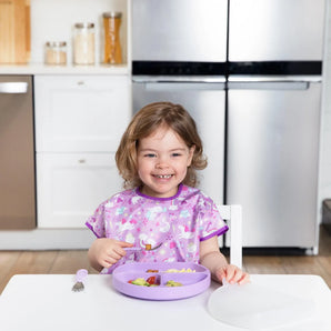 A smiling toddler in a purple bib sits at a white table with a Bumkins Silicone Grip Dish with Lid (3 Section): Lavender in a kitchen.
