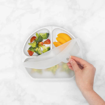 Opening a Silicone Grip Dish with Lid by Bumkins, revealing toddler portions of broccoli, tomatoes, and mango on a Marble plate.