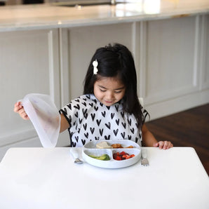 At a white table, a girl in a heart-print bib lifts the lid from a Bumkins Silicone Grip Dish with Lid (3 Section): Marble with snacks.