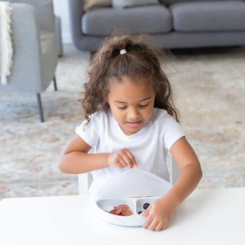 A curly-haired girl in a white shirt opens Bumkins Silicone Grip Dish with Lid (3 Section): Marble while sitting at a table, sofa behind.
