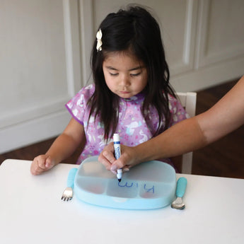 A young girl sits at a table as an adult writes with a marker on a Bumkins Silicone Stretch Lid for Grip Dish 5 Section, Grip Tray.