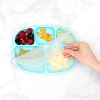 A hand lifts Bumkins Silicone Stretch Lid for Grip Dish 5 Section, Grip Tray, revealing a blue plate with food on a white marble surface.