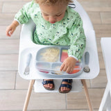 In a high chair, a toddler in a green animal-print bib lifts a Bumkins Silicone Stretch Lid for Grip Dish 5 Section, Grip Tray on a tray.