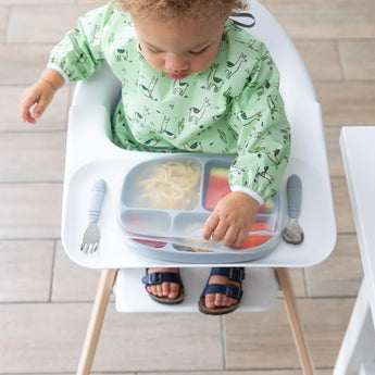 In a high chair, a toddler in a green animal-print bib lifts a Bumkins Silicone Stretch Lid for Grip Dish 5 Section, Grip Tray on a tray.
