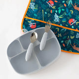 Gray Silicone Grip Dish by Bumkins, featuring a lid and 5 sections, beside a spoon and fork on an animal-themed bib. Food-safe silicone.