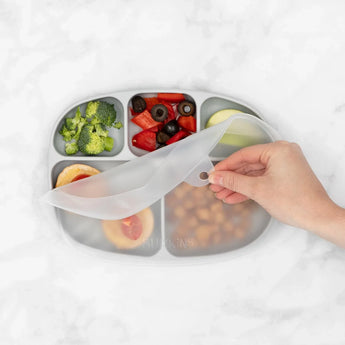 A hand covers a Bumkins Silicone Grip Dish with Lid (5 Section): Gray, filled with broccoli, olives, and more.