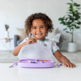 A child smiles, lifting a Bumkins Silicone Stretch Lid for Grip Dish 5 Section, Grip Tray on top of a purple plate in a bright room.
