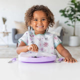 A smiling child with a bib pulls the tab of a Bumkins Silicone Stretch Lid for Grip Dish 5 Section, Grip Tray at a table in a bright room.