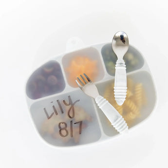 A fork and spoon rest on a Bumkins Silicone Stretch Lid for Grip Dish 5 Section, Grip Tray, covering a plate of food on a white background.