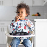 A toddler in a bib enjoys snacks from a dish while in a high chair, all from Bumkins Disney Little Ones Gift Bundle: Mickey Mouse Classic.