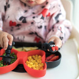 A child in a bib enjoys veggies and fruit from a dish with utensils, all from Bumkins Disney Little Ones Gift Bundle: Minnie Mouse Classic.