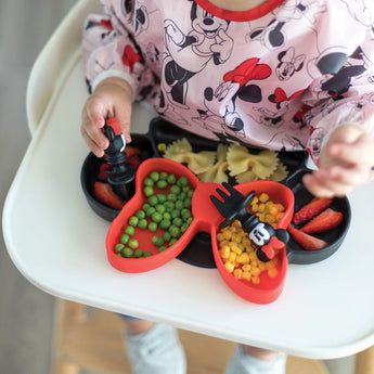 A child in a high chair in a bib enjoys food from a dish with utensils, all from Bumkins Disney Little Ones Gift Bundle: Minnie Mouse Classic.