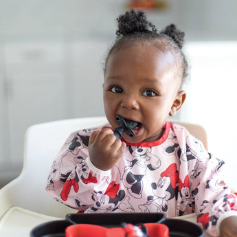 A baby in a high chair curiously chews a utensil while wearing a bib, all from Bumkins Disney Little Ones Gift Bundle: Minnie Mouse Classic.