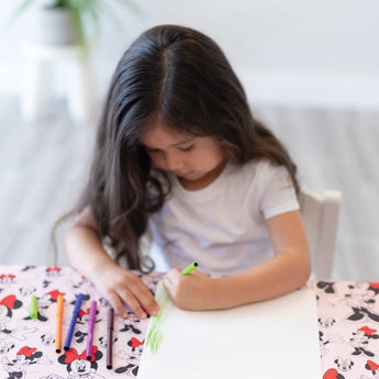 A young girl with long hair draws at a table covered in a splat mat from Bumkins Disney Little Ones Gift Bundle: Minnie Mouse Classic.