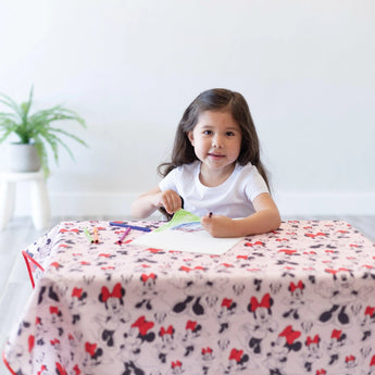 A girl doodles at a table covered in a splat mat from Bumkins Disney Little Ones Gift Bundle: Minnie Mouse Classic with a plant nearby.