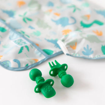 Close-up of a dinosaur pattern bib and silicone utensils from Bumkins Ultimate Gift Set - Dinosaurs & Blue Tropic on a white background.