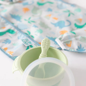 Close-up of a dinosaur-themed bib and bowl with spoon and lid from Bumkins Ultimate Gift Set - Dinosaurs & Blue Tropic on a white background.