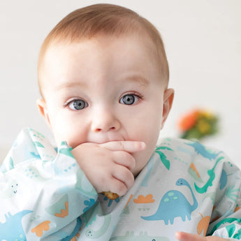 A baby in a dinosaur-print bib from Bumkins Ultimate Gift Set - Dinosaurs & Blue Tropic in a bright room.