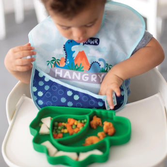 A child in a bib peeks into their pocket as they eat from a dinosaur dish, all from Bumkins Ultimate Gift Set - Dinosaurs & Blue Tropic.