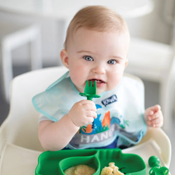 A baby in a high chair in a bib, uses utensils to eat from a dinosaur dish, all from Bumkins Ultimate Gift Set - Dinosaurs & Blue Tropic.