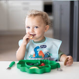 A curly-haired toddler in a bib eats at a table from a dinosaur-shaped plate, all from Bumkins Ultimate Gift Set - Dinosaurs & Blue Tropic.