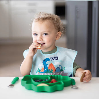 A curly-haired toddler in a bib eats at a table from a dinosaur-shaped plate, all from Bumkins Ultimate Gift Set - Dinosaurs & Blue Tropic.