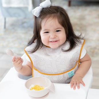 A toddler with a bow in a high chair wears a beige bib and eats from a beige bowl from Bumkins Ultimate Gift Bundle, Desert Boho.