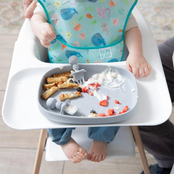 A baby in a high chair wearing a blue bib relishes yogurt and snacks from Bumkins Grip Tray Essential 4-Piece Mealtime Set: Gray.