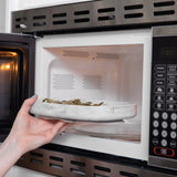 A hand places a Bumkins Silicone Grip Tray: Marble filled with food into an open microwave.