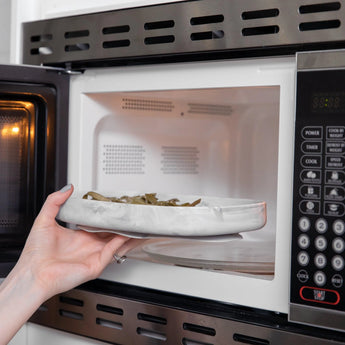 A hand places a Bumkins Silicone Grip Tray: Marble filled with food into an open microwave.