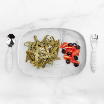 Bumkins Silicone Grip Tray: Marble holds spinach pasta, cherry tomatoes, and olives, alongside a spoon and fork on a marble background.