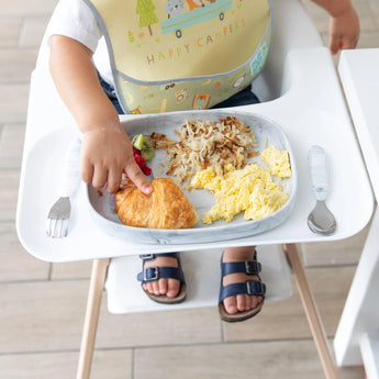 A toddler in a high chair enjoys breakfast from a Bumkins Silicone Grip Tray: Marble, with scrambled eggs, hash browns, and a croissant.