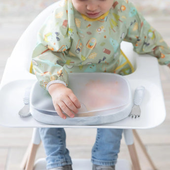 A toddler in a high chair reaches for the tab of a Bumkins Silicone Stretch Lid for Grip Dish 5 Section, Grip Tray covering a plate.