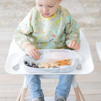A toddler in a high chair removes a Bumkins Silicone Stretch Lid for Grip Dish 5 Section, Grip Tray from a tray with berries and sandwiches.