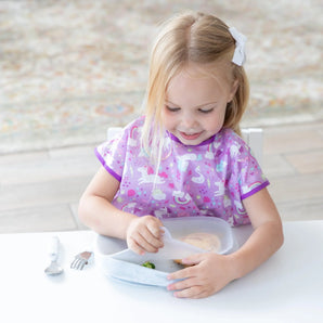 A young blonde child in a purple bib sits at a white table, removing a lid from a Bumkins Silicone Grip Tray: Marble.