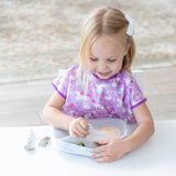 A young blonde child in a purple bib sits at a white table, removing a lid from Bumkins Silicone Grip Tray: Marble with utensils nearby.